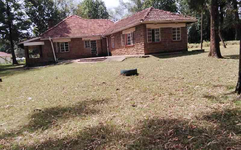 A single-storey stone house with a tiled roof, surrounded by a lawn and mature trees.