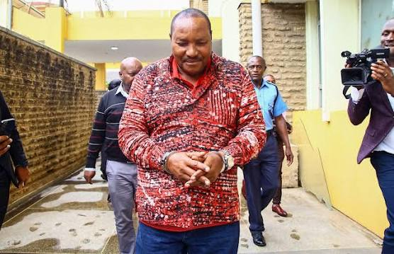 Ferdinand Waititu walking outdoors in a red patterned shirt, accompanied by other people and a camera operator.