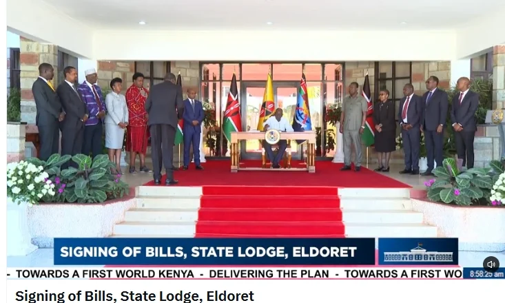 A televised bill-signing ceremony at State Lodge in Eldoret, with officials standing beside the signing table.